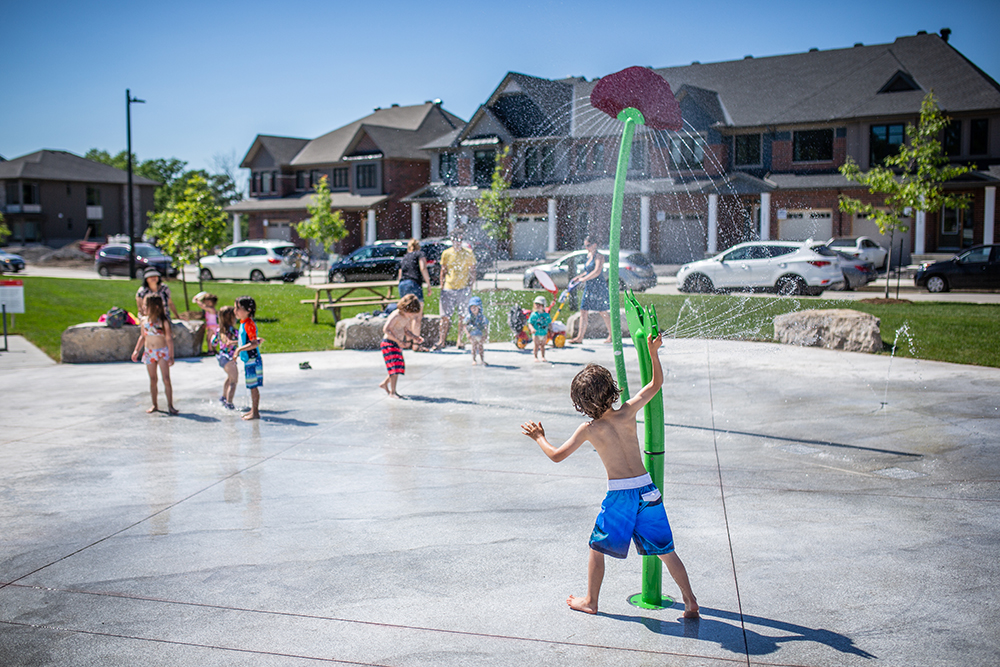 A small boy playing at an outdoor water playground. Houses line the background.